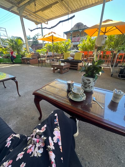 The image depicts an outdoor seating area of a cafe, likely in Bellingen, Australia, characterized by its relaxed ambiance and natural surroundings. In the foreground, a person's legs are visible, clad in black trousers with a floral pattern and white sneakers, seated at a dark wood coffee table with a glass top. On the table, there's a coffee cup with a strainer, a small metal pot, a spoon, and a bucket labeled "Habitat Coffee House" containing what appear to be sugar packets. A potted plant sits on the table, adding a touch of greenery.
The cafe's outdoor area features a covered patio with a metal roof and exposed beams, providing shade from the sun. Several tables and chairs are arranged around the space, with orange chairs visible in the background. Lush green plants and palm trees are scattered throughout the area, contributing to a tropical feel. In the middle ground, a woman is seated on a green outdoor sofa, reading a book. Further back, other patrons are seen at tables, some engaged with laptops, suggesting a common gathering spot. The background reveals a building with a distinctive architectural style, possibly a heritage building, with balconies and awnings. The presence of orange umbrellas and the overall outdoor setting indicate a warm, possibly sunny day, consistent with the climate of Bellingen. The scene conveys a sense of casual leisure and community, where people can relax, enjoy refreshments, and socialize.