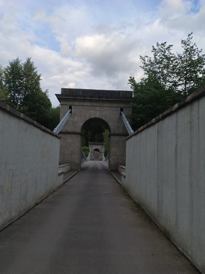 The image depicts a view looking down a paved pathway flanked by two substantial, light-colored concrete walls. The pathway leads towards an imposing stone archway, which appears to be part of a suspension bridge structure. Beyond the archway, a second, similar archway is visible in the distance, also part of the bridge. Cables from the suspension bridge are visible, extending upwards from the pathway and disappearing into the archway's structure. Trees line the background, with a cloudy sky above. The scene suggests a park or historical site. This is the Slavkovice suspension bridge, located in Slavkovice, Czechia.