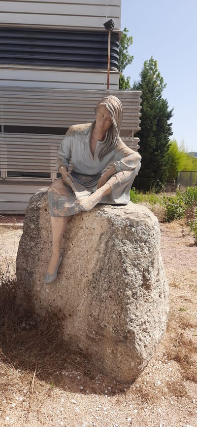 A statue of a woman reading is seated on a large boulder in Vacarisses, Spain. The statue, made of a light-colored material, depicts a woman with long hair, wearing a long-sleeved tunic and a skirt. She is leaning forward, her head bowed, as if engrossed in a book or document she holds in her hands. Her legs are crossed, with one foot resting on the boulder. The boulder itself is rough and uneven, with a lighter, speckled surface. Behind the boulder and statue, a building with horizontal grey louvers is visible. In the background, there are trees, green foliage, and a clear blue sky. The ground in front of the boulder is covered in dry grass and small gravel. The overall scene suggests an outdoor setting, possibly a park or public space.
