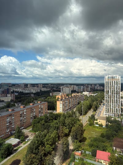 The image captures an aerial view of a cityscape under a cloudy sky, with a focus on residential buildings and abundant greenery. The sky is dominated by large, dark, and fluffy clouds, with patches of blue sky and lighter clouds visible towards the horizon.
In the foreground, several multi-story brick apartment buildings are prominent. One such building, a long, reddish-brown structure, occupies the lower left quadrant. It features numerous balconies and windows. To its right, a modern, tall, white building with a distinctive pattern of green and white windows stands out, extending vertically through the middle ground and into the distance.
The cityscape is interspersed with dense clusters of trees, forming green belts between the buildings and lining a road that snakes through the scene. The road, visible in the middle ground, is lined with parked cars. Further in the distance, a sprawling urban landscape of apartment blocks and other structures extends to the horizon.
A small, yellow, gabled building is situated in a grassy area to the right of the road, near the tall white building. In the lower right corner, part of a red-roofed building is visible, suggesting a more industrial or utility area. A sidewalk and a small, kiosk-like structure are present in the lower left quadrant.
The scene appears to be an urban residential area, characterized by a mix of older and newer architecture, with significant presence of green spaces. The overall impression is one of a densely populated city district with a balance between built environment and nature. The location is identified as Verkhnii Liuk, Russia, based on the prompt. There are no visible people or specific actions captured in the image.