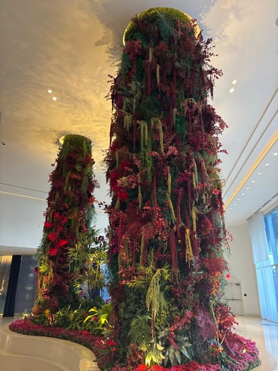 The scene depicts an expansive interior space in Winchester, United States of America, featuring two prominent, floor-to-ceiling columnar structures entirely covered in dense botanical arrangements. The larger column, positioned centrally and to the right, extends to the ceiling and is adorned with a rich mixture of deep green, crimson, and maroon foliage, interspersed with numerous hanging strands of lighter green and yellowish plant material. A second, shorter column is partially visible in the left background, exhibiting a similar dense botanical covering dominated by green and red hues. Both columns originate from raised, curvilinear planters at their bases, which are also populated with similar plant species. A smooth, light-colored curved architectural element is visible in the lower-left foreground. The overhead ceiling is light-toned and features recessed spotlights casting warm illumination, with shadows of the botanical structures distinctly visible. The floor is light-colored and reflective. In the background on the right, large windows draped with white sheer curtains are visible, alongside a white exit door. The background on the left includes a darker wall with subtle geometric patterns. No individuals are present within the frame.