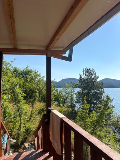 The image shows a view from a wooden porch overlooking a body of water and hills. A towel and dark clothing are hanging on the wooden railing of the porch. There is lush greenery and trees surrounding the porch, and a clear blue sky above. In the background, there are rolling hills covered in trees, and a calm body of water. The scene appears to be in a natural, possibly rural, setting. The location is specified as Zvanika, Bulgaria.