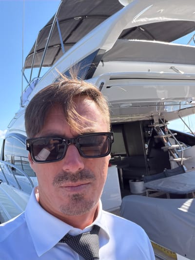 A medium shot captures a man wearing sunglasses, a white collared shirt, and a black and white polka dot tie. He is standing outdoors, with a large white yacht behind him. The sky is blue and clear, suggesting a sunny day. The man's sunglasses reflect a portion of the yacht. The image appears to be taken in a harbor or marina. The Village of Sag Harbor is a village in Suffolk County, New York, United States. It is located in the town of Southampton and the town of East Hampton.