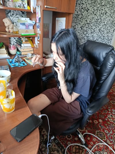 The original image shows a young woman with long black hair sitting at a wooden desk in a room. She is wearing a dark t-shirt and brown shorts. Her left hand is raised, and she appears to be holding something with long, pointed fingernails. On the desk, there is a black smartphone plugged into a charger, two mugs with yellow flower designs, and various other items including books and what appear to be photos. The background includes shelves filled with books and decorations, and a patterned wallpaper. The floor is covered with a red, patterned carpet. The image does not provide any specific information about the location in the city of Kostopil, Ukraine.