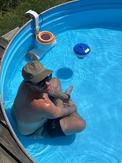A medium shot from above shows a man sitting in a circular, above-ground swimming pool filled with blue water. He is wearing a green baseball cap, sunglasses, and dark swim trunks. His arms are crossed, and he appears relaxed. The pool has a metal rim and a wooden deck surrounding it. In the background, there is green grass and a pool skimmer attached to the rim, with a blue chemical dispenser floating in the water. The sun is shining, casting shadows on the water. The image's metadata indicates the location as San Lorenzo de Tormes, Spain.