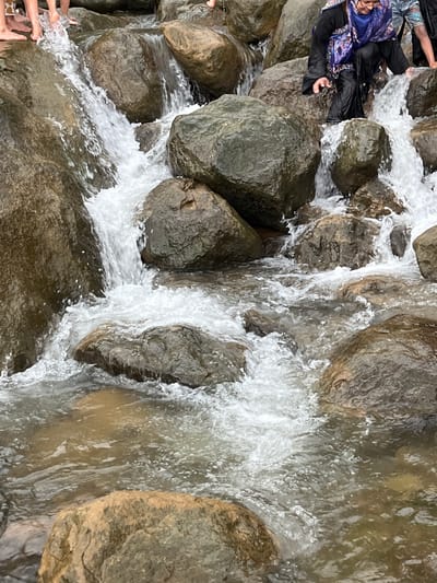 In Jaflong, Bangladesh, the image captures a close-up view of a shallow river with clear, flowing water cascading over numerous rocks. Several people are present, some wading in the water with their feet visible, while others, including a woman in traditional attire, are navigating the rocky terrain near the water's edge. The environment is characterized by a rocky riverbed and surrounding boulders, with the water appearing somewhat murky. The scene depicts a natural setting with human interaction, suggesting activities such as crossing the river or enjoying the water.