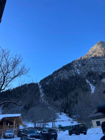 This image, originally captured in portrait orientation and rotated 90 degrees counter-clockwise, depicts a bright winter scene in Courmayeur, Italy. The vast, clear blue sky dominates the upper left portion of the frame, indicating good weather.

A large, rugged mountain, partially covered in snow and rock, rises prominently on the right, bathed in strong sunlight. Below the mountain, a dense evergreen forest blankets the slopes, also showing signs of snow.

In the midground and foreground, on the bottom right, is a snowy parking area occupied by several vehicles, including a large, dark SUV resembling a Hummer H3 and other passenger cars. To the far right, a building with a dark roof and light walls is visible, possibly part of a resort. Closer to the foreground, a wooden structure with a snow-covered roof and exposed beams appears to be under construction or recently built. A prominent bare-branched deciduous tree stands in the lower right, adding to the winter aesthetic. Faint structures resembling ski lift components can be discerned among the trees in the midground.

The scene is set during daylight, likely morning or early afternoon, given the clear sky and bright illumination on the mountain and foreground elements. The presence of snow, ski lift elements, and resort-style buildings suggests a winter sports destination. No people or visible text are discernible in the image.