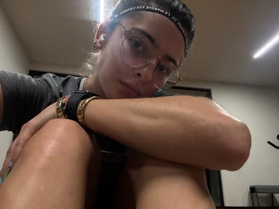 An individual is depicted in a close-up, low-angle indoor photograph taken in Komchén, Mexico. The subject, a young adult with light skin and dark hair pulled back, wears a grey t-shirt, clear-rimmed round glasses, and a black headband featuring the text "LA MARQUE AUX 3 BANDES /// THE BRAND WITH THE 3 STRIPES." White wireless earbuds are present in both ears. The left wrist displays an Apple Watch with a black band and multiple gold-colored bracelets, one with a red charm. The individual's visible fingernails exhibit blue polish. They are seated, leaning forward with the head or chin resting on the right arm, while the left arm is bent, hands resting on the upper leg. The skin appears slightly shiny, suggesting recent physical activity. The background consists of a light-colored wall, with a dark rectangular object (possibly a doorframe) behind the subject's left shoulder. To the right, blurred, are components of exercise equipment, including a black machine and a grey bench. Overhead linear fluorescent lighting illuminates the scene from the top center.