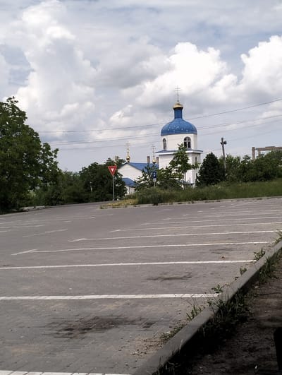 The image shows a parking lot with white lane markings in the foreground, leading towards a street and a church in the background. The church has a white facade and a prominent blue dome with golden crosses. Trees and green foliage are visible on either side of the church, and a yield sign is positioned near the road. The sky is overcast with scattered white clouds. The environment appears to be a semi-urban or rural setting. This scene is likely located in Șoldănești, Moldova, given the context.