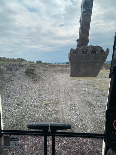 The image, taken from the perspective of an excavator operator, shows a view of a gravel pit. The excavator's bucket, filled with dirt and rocks, is prominently positioned in the upper center of the frame, obscuring the sky. The terrain is uneven and covered with small to medium-sized stones and gravel. Tire tracks are visible on a dirt path leading into the pit. In the foreground, the control levers of the excavator can be seen. The background reveals a distant treeline and hills under a cloudy sky. The scene appears to be an active construction or excavation site. There is no visible indication of the specific location in Zmeiskaya, Russia.