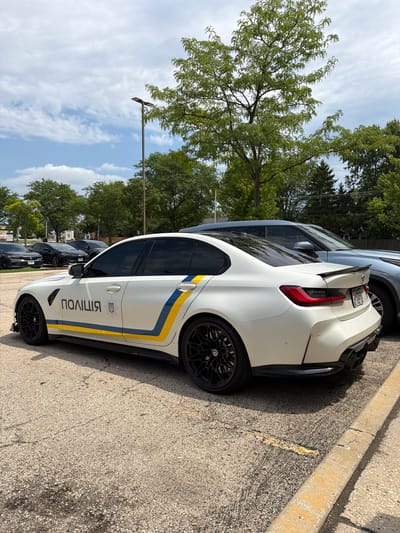 A white BMW M3 sedan, marked with "ПОЛІЦІЯ" in black lettering and a Ukrainian trident emblem, is parked in a lot in Buffalo Grove, Illinois. The car features blue and yellow stripes along its side, a black rear spoiler, and black wheels. Behind the BMW, a light blue SUV is parked. The scene is set outdoors on a paved lot with trees and a cloudy sky in the background.