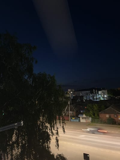 The image depicts a nighttime urban scene in Raduga, Russia, viewed from an elevated perspective.

In the immediate foreground, obscuring the lower left, are dark, dense tree branches with small, pointed leaves, likely from a deciduous tree. A metallic, cylindrical railing or bar extends diagonally from the lower-left edge towards the center-bottom.

Below, a multi-lane asphalt road runs horizontally across the frame, brightly illuminated by unseen streetlights, creating a somewhat reflective surface. White dashed lines clearly mark the lanes. A single vehicle is captured in motion on the road, rendered as an elongated blur due to a long exposure. Its headlights appear as bright white streaks moving left-to-right, while its taillights are distinct red streaks, indicating a continuous rightward trajectory. A small, dark, rectangular object, possibly a sign or sensor, is positioned on the bottom right edge of the frame.

To the right of the road, several low-rise buildings and associated fences are visible. These include a brick-colored house with a dark, ridged roof and partially legible signs on its facade, and a smaller, lighter-colored structure with a blue gate and fence in front. Surrounding vegetation, including trees and shrubs, is present around these structures.

In the mid-background, a cluster of multi-story buildings, some appearing residential or commercial, are partially lit by internal and external lighting, their forms silhouetted against the dark sky. One prominent, light-colored building features multiple windows and what appear to be ventilation shafts or pipes on its exterior. A distant horizon line shows a faint, diffuse glow, suggestive of ambient light pollution from the city.

The dark, deep blue night sky occupies the upper portion of the frame. Two prominent, broad, diffuse streaks of light, oriented diagonally from the upper left to the upper right, are visible across the sky. These appear to be reflections on the viewing surface (e.g., a window) rather than atmospheric phenomena. A few faint, scattered point lights, indicative of distant stars, are sparsely visible in the darker areas of the sky. No human figures are discernible in the scene.