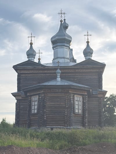 An exterior, eye-level, medium shot captures a Russian Orthodox wooden church with multiple onion domes topped with crosses. The church, constructed of dark, weathered logs, features a prominent octagonal section with a multi-paned window. The sky above is overcast with scattered clouds. The foreground shows tall green grass and a patch of dark soil. The location is the city of Cherdyn, Russia, specifically the Church of the Transfiguration. No people are visible in the image.