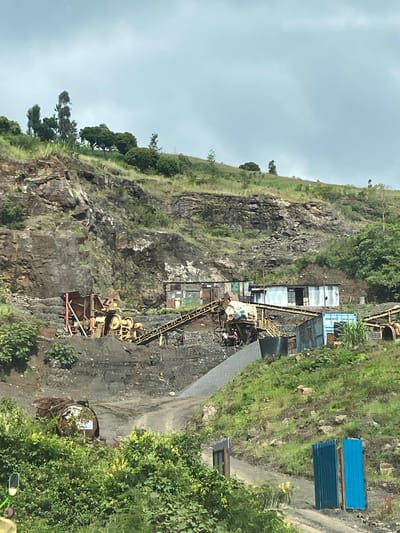 This image captures an active quarry or rock-crushing site nestled against a steep, rocky hillside under a cloudy sky. The scene is situated in Kinyona ward, Kenya.
The prominent feature is the rugged hillside, characterized by exposed grey and brown rock formations. Patches of green grass, various trees, and bushes grow on and around the rock, indicating some natural vegetation amidst the industrial activity.
At the base and mid-section of the hillside, an array of heavy industrial machinery, predominantly yellow, is visible. This includes a large rock crusher, an extensive system of conveyor belts extending up the slope, and other processing equipment used for material extraction and sorting. Piles of dark, processed aggregate are scattered throughout the operational area.
Several utilitarian structures, likely temporary offices or workshops, are present. These are constructed from corrugated metal sheets, with some appearing grey/silver and others a distinct blue. Two prominent blue corrugated panels are stacked vertically on the right side of the image, near a dirt path that winds through the site. A large, dark cylindrical object, possibly a tank or container, is also visible near the machinery.
A few indistinct figures, presumably workers, can be observed near the processing equipment, suggesting ongoing operations. The diffused light indicates a cloudy daytime setting. The scene portrays a busy industrial landscape carved into a natural environment. No legible text is visible in the image.