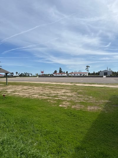 This image depicts an aerodrome or airfield located in Praia do Ribatejo, Portugal, under a clear daylight sky. The foreground features a vibrant green grassy area transitioning to patches of sparse, lighter-colored vegetation and bare earth. A small, reddish-brown rectangular marker is visible on the left within the green grass. The midground is dominated by a wide, dark-colored paved surface, resembling an asphalt runway or taxiway, running horizontally across the frame with faint white markings. In the background, a series of single-story white buildings with reddish-brown roofs are visible, separated from the paved area by a low, dark barrier. To the far left, a red metal tower structure, possibly a control tower, stands prominently. Further right, a larger grey metal building with a dark roof, likely a hangar, is present. Numerous pine-like evergreen trees are interspersed among and behind the buildings, forming a green line on the horizon. The sky is light blue with scattered cirrus clouds and two distinct, parallel white contrails in the upper left quadrant. No people, vehicles, or active aircraft are visible.