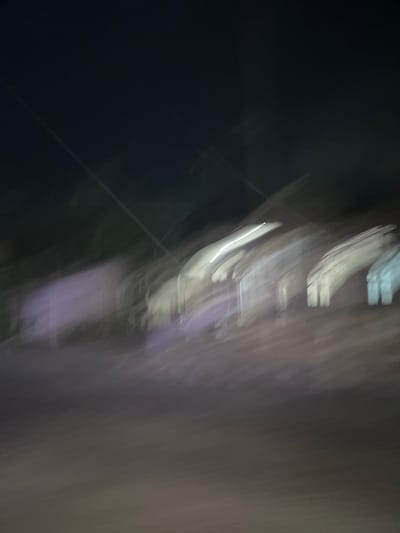 This media file is a blurry, abstract photograph taken at night, possibly in Landuh, Indonesia. The image appears to capture a scene with artificial lights, creating streaking effects due to motion blur. On the right side, there are vertical streaks of white and light purple, suggesting illuminated objects or structures. The left side is predominantly dark, with some faint, lighter lines that could be reflections or distant lights. The overall impression is one of movement and low light conditions, making it difficult to discern specific subjects or a clear setting. There is no discernible text in the image. The abstract nature of the photograph prevents any detailed observation of activity, emotions, or specific location cues.