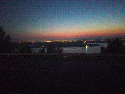 The image depicts a cityscape at dusk, likely Veselyy, Russia, with a blend of natural and artificial lights. The sky transitions from a deep blue at the top to hues of orange and pink near the horizon, indicating the setting sun. Below the sky, a line of city lights glows, suggesting the presence of buildings and infrastructure. A body of water, possibly a river or lake, is visible in the middle ground, reflecting some of the artificial lights. The foreground is dark and indistinct, with what appear to be trees or other vegetation silhouetted against the brighter background. The image is grainy, suggesting it was taken in low light conditions. There are no visible people or animals in the frame, and the focus is on the overall atmospheric landscape.
