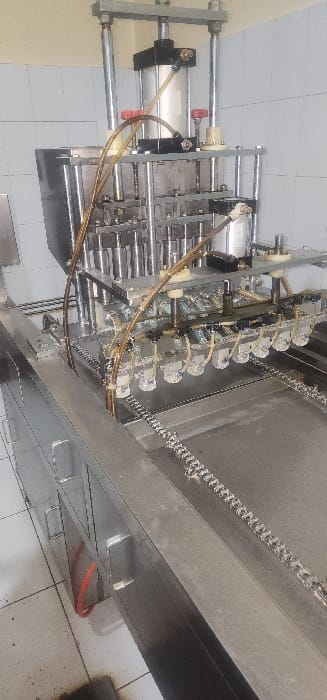 This image captures a close-up view of an industrial filling or dispensing machine within an indoor setting. The primary subject is the complex arrangement of metallic machine components designed for automated production.

The machine features multiple pneumatic cylinders, two of which are prominently visible on the left, mounted vertically within a robust metal frame. These cylinders are connected by clear and red air hoses, indicating they are operated by compressed air. Further to the right, a series of at least ten identical dispensing nozzles or spouts are arranged in a linear fashion. Each nozzle appears to have individual control mechanisms, possibly valves or smaller actuators, connected by additional hoses. This configuration strongly suggests a multi-head system for accurately filling containers or dispensing materials.

Adjacent to the dispensing heads, a metal conveyor system with a visible chain mechanism is present on the right side of the frame. This chain conveyor is likely used to transport items, such as bottles or packages, through the filling station. The machine's structure is predominantly made of clean, polished, or brushed metal, suggesting durability and suitability for an industrial environment.

The setting appears to be a manufacturing or processing facility, indicated by the tiled floor visible beneath and around the machinery. No specific activity is being performed at the moment the photo was taken, but the machine's design clearly implies its function in an automated production line. The equipment appears well-maintained and clean. There is no visible text or branding on any part of the machinery.