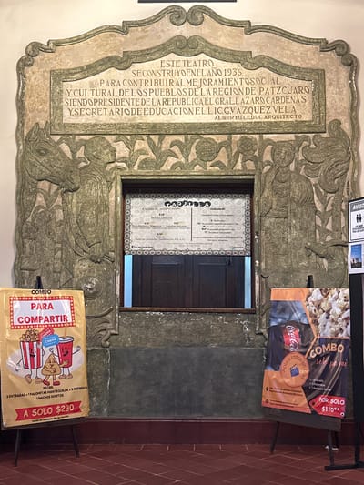 This image captures the entrance area of the Teatro Emperador Caltzontzin, located in Plaza Gertrudis Bocanegra S/N, Centro, Pátzcuaro, Mexico. The focal point is a large, elaborate sculpted stone edifice framing a closed ticket booth.
The upper section of the stone structure features a carved plaque with Spanish text. It states that the theater was constructed in 1936 to contribute to the social and cultural improvement of the Pátzcuaro region, under President Lázaro Cárdenas and Secretary of Education Lic. G. Vázquez Vela, with Alberto Leduc as the architect.
Below the plaque, two intricate stone relief carvings flank the rectangular ticket window. The carving on the left depicts a stylized male figure standing beside a horse's head and neck, holding a circular object, amidst foliage. The carving on the right shows a female figure with prominent headwear or hair, holding an item, with foliage and a serpentine or aquatic creature at her base. The ticket window itself is dark, featuring two closed wooden panels. Inside the frame above the window, a "TAQUILLA" (Ticket Office) sign is visible, detailing "Cuotas de recuperación" (Recovery fees) at $35 for general, students/seniors, and children, along with RTC film classifications.
The floor is covered in reddish-brown terracotta tiles. Two advertising posters on black easels stand in the foreground. The yellow poster on the left advertises a "COMBO PARA COMPARTIR" (Combo to Share) for $230, illustrated with cartoon popcorn, soda, and nachos, listing two tickets, butter popcorn, two sodas, and Doritos nachos. The dark poster on the right promotes a "COMBO solo pa' mi" (Combo just for me) for $110, showing popcorn and a Coca-Cola logo, detailing one ticket, popcorn, and a 600ml soda. A partially visible white sign on the far right displays "AVISO" and restroom symbols. No individuals are present in the scene.