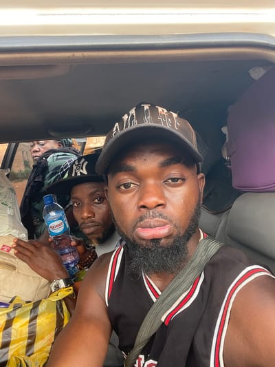 Three men are in a vehicle. The first man, in the foreground, wears a black and red basketball jersey and a hat. A seatbelt is across his chest. He is sticking his tongue out. The second man holds a water bottle. The third man is in the background. A yellow and black striped bag and luggage are also visible. Based on other images from the same source, the location is in Otukpa, Nigeria.