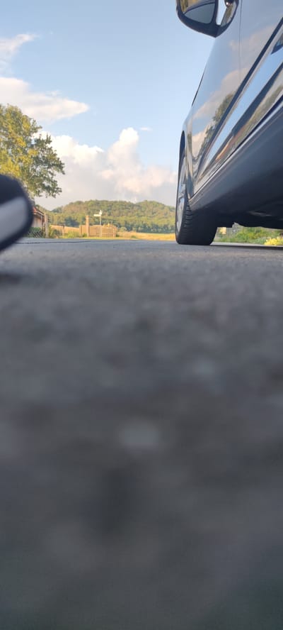 The image captures a low-angle view of a road with a car's rear tire and side visible on the right. The car appears to be parked or moving slowly on an asphalt road. In the background, a tree with green foliage is on the left, and a fence runs horizontally across the scene. Further back, rolling green hills with scattered trees form the landscape under a bright blue sky with scattered white clouds. There are no people visible in the image. The scene is likely set in a rural or semi-rural area, possibly on the outskirts of Liebenburg, Germany, given the presence of a road, natural landscape, and a car.