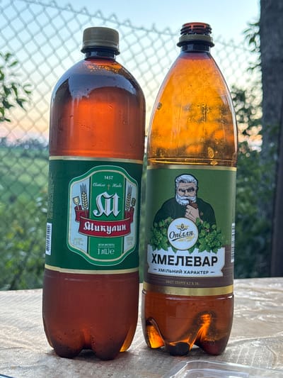 The image shows two large plastic bottles of beer on a table outdoors. The bottle on the left has a green label with a wheat motif and the text "1457 CIMILE + HUEBE" and "ШКУЛИН" in Cyrillic. The bottle on the right has a green label featuring an illustration of an old man with a beard and pipe, and the text "Опілля", "ХМЕЛЕВАР" and "ХМІЛЬНИЙ ХАРАКТЕР" in Cyrillic. The background shows a wire fence and greenery, suggesting an outdoor setting. The presence of condensation on the bottles indicates they are cold. The lighting suggests it is late afternoon or early evening. The table surface is textured and appears to be made of a woven material. A clear plastic container is partially visible in the bottom right corner of the image. The scene is set in the village of Krasivka, Ukraine.