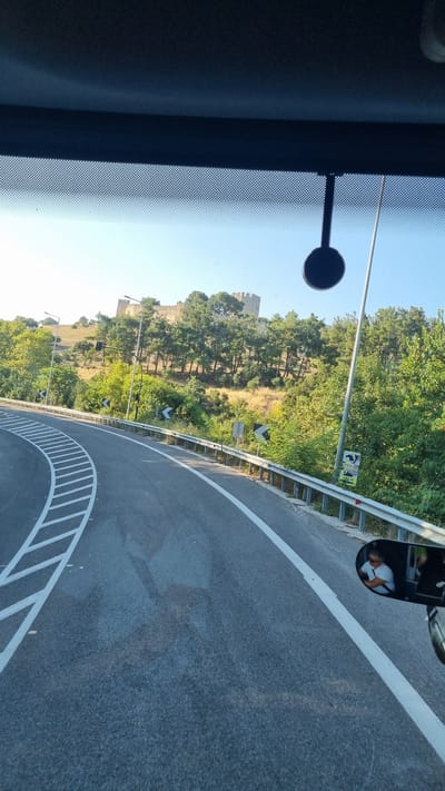 The image captures a view from inside a vehicle looking out through the windshield at a road curving to the left, leading towards a hillside with a stone fortress or castle structure on its summit. The fortress, with its crenellated walls, is situated amidst dense green trees and a sparse landscape under a clear blue sky. The road is asphalt, marked with white lane lines and diagonal white markings indicating a change in lane. A metal guardrail runs along the right side of the road, with several black and white directional signs attached to it. There is a lamppost next to the road, and a black circular object, possibly an air freshener, is hanging from the vehicle's interior. In the side mirror, a person wearing a white t-shirt is visible, appearing to be the driver, and another person is partially visible behind them. The scene suggests a scenic drive on a sunny day in or near Neos Panteleimonas, Greece, with the historical fortress serving as a prominent landmark.