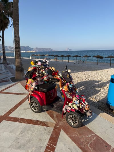 The image shows a red motorized scooter heavily decorated with numerous stuffed animals. The scooter is parked on a marble-tiled promenade adjacent to a pebble beach. In the background, the Mediterranean Sea is visible with several beach umbrellas and lounge chairs set up on the sand. Palm trees line the promenade, and distant mountains form the horizon. The scene is set in l'Alfàs del Pi, Spain, a coastal town known for its beaches. There are no people actively interacting with the scooter in the image.