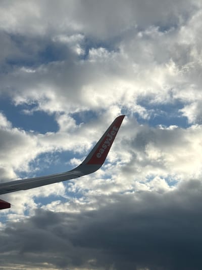 The image depicts the tail fin of an EasyJet airplane against a backdrop of a cloudy sky. The tail fin is red with the EasyJet logo in white. The clouds range from light and puffy to dark and ominous. The location in the city of Berg, Luxembourg, is not apparent from the visual information provided in the image.
