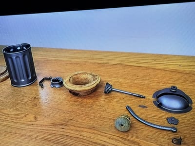 An assortment of disassembled parts from a coffee grinder are laid out on a wooden table. The parts include a dark grey cylindrical hopper with vertical grooves, a metallic clamp, a wooden bowl-shaped component, a metal shaft with a grinding burr, a small flat metal piece, a wooden knob, a curved metal handle with perforations, a small gear-like metal piece, and a dark grey domed lid with a handle. The background consists of a plain light blue wall. The scene is located in Castillo Bajo-conejito, Spain.