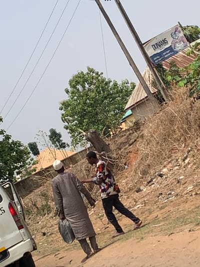 The scene depicts a street-level view in the city of Tataura, Nigeria, with a white vehicle partially visible at the bottom of the frame. Two individuals are present. One is wearing a long grey robe and a white cap, and the other is wearing a patterned shirt and dark pants. They are standing in close proximity, possibly interacting, near a dirt road and dry vegetation. A sign with the word "Dunamis" is visible in the background, attached to a utility pole. Buildings with corrugated roofs are also visible, partially obscured by trees and foliage. Power lines extend across the upper left portion of the frame.