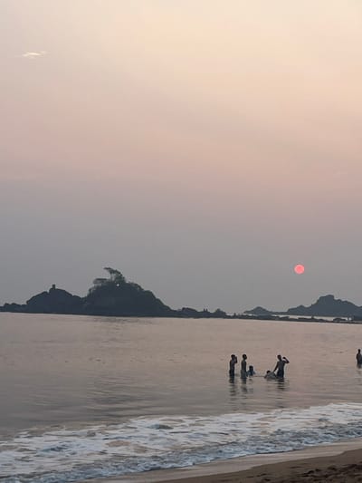 This image depicts a coastal scene at twilight, consistent with a sunset, on a beach in Gokarna, India. The sky exhibits a gradient from muted pink to pale orange, with a distinct, spherical red sun positioned in the upper right quadrant. A singular, small white cloud is visible in the upper left.

The foreground features a narrow strip of wet sand with a foamy white surf line. The main body of water is a calm sea, reflecting the sky's hues. Seven human figures are present within the shallow water, roughly waist to chest deep. On the left-center, a group of four individuals includes an adult-sized figure standing near a smaller, partially submerged individual. In the mid-center, two figures stand, one with a raised right arm. A seventh, solitary figure is discernible further to the right. All figures are silhouetted against the water and sky.

In the background, a series of dark, rocky landmasses form a silhouetted horizon. The most prominent landmass, positioned left-center, features a cluster of trees at its crest and a distinct, slender, man-made structure on its left flank. Other smaller rocky outcrops are visible further right along the horizon.