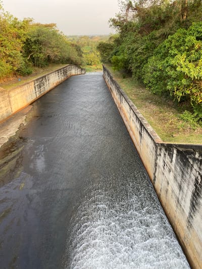 This is a media file captured from a high vantage point, looking down a concrete spillway at a dam. The spillway is filled with flowing water, creating a rippling effect as it moves downstream. The water is a dark gray, with the whitewater from the spillway's edge contrasting sharply. The spillway walls are made of concrete, with a textured surface and visible signs of wear and staining, suggesting age and exposure to the elements. Green vegetation, including trees and grass, lines the sides of the spillway, indicating a natural, possibly rural, setting. The sky in the background appears overcast and hazy, suggesting a potentially cool or damp day, possibly early morning or late afternoon given the soft, diffused light. There is no visible text within the image. The overall impression is of a functional piece of infrastructure within a natural landscape, with the dynamic movement of water being the primary focus.