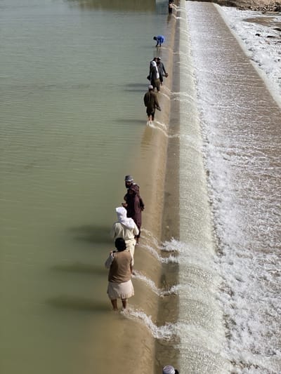 A high-angle, long-shot photograph shows a line of several men crossing a dam in what appears to be a river. The location is in the city of Kim, Afghanistan. The water is a muddy green color and flows from left to right. The men are wading through the water as they walk along the dam's edge, some are dressed in traditional Afghan attire, including long robes and head coverings. The sky is not visible, but the light suggests a bright, sunny day. The dam is made of concrete and has water flowing over its edge.