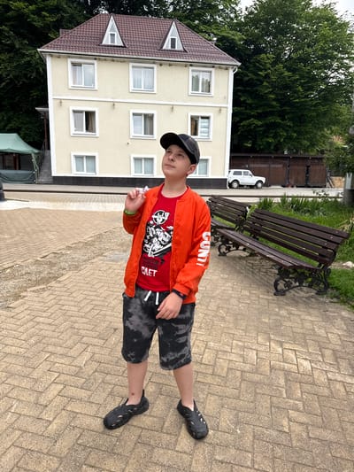The image shows a young boy standing on a paved walkway in front of a building in Guamka, Russia. The boy is wearing a red t-shirt with a graphic print, an orange bomber jacket, grey tie-dye shorts, and black sandals. He is looking upwards and holding a small white object in his right hand. Behind him is a two-story building with a brown roof and several windows. The building is surrounded by lush green trees. In the background, there is a white car parked next to a wooden fence. Two park benches are visible to the right of the boy. The ground is paved with light brown rectangular bricks laid in a herringbone pattern.