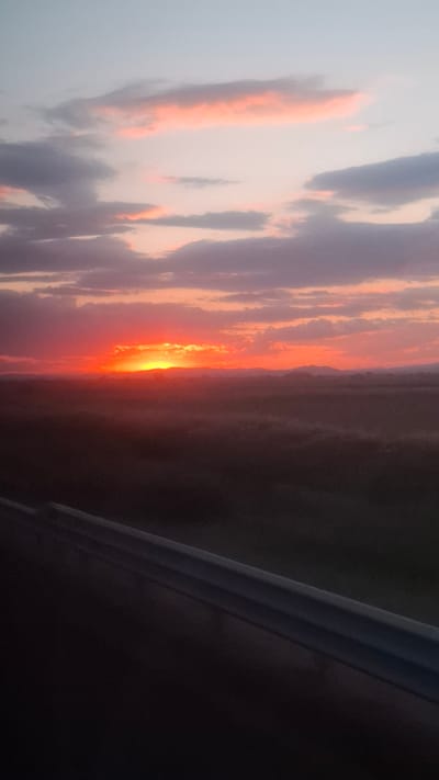 This is a vertical, eye-level, medium shot photograph of a sunset over a rural landscape. The sun, a vibrant orange ball, is just above the horizon, casting a fiery glow across the sky. Clouds, tinged with pink and purple, are scattered across the horizon, some dark and foreboding, others light and wispy. Below the sky, the landscape is a dark expanse of fields and rolling hills, partially obscured by a light mist. In the foreground, a dark, metallic barrier runs diagonally across the bottom of the frame, suggesting the vantage point is from a bridge or elevated structure. The overall mood is one of dramatic beauty, with the intensity of the sunset contrasting with the subdued tones of the landscape. The location is the village of Tsalapitsa, Bulgaria.