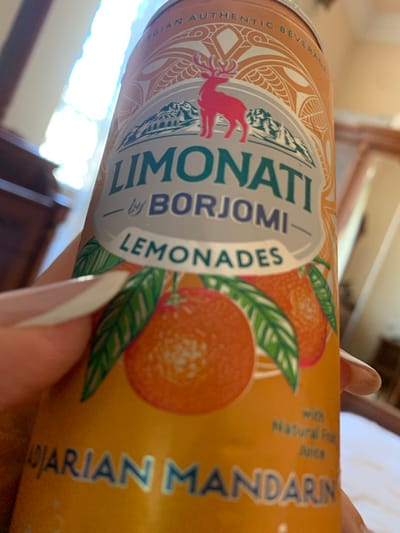 A close-up, slightly angled shot shows a hand holding a bright orange beverage can. The can features a stylized design with a pink deer, mountains, and stylized fruit, possibly oranges or mandarins, with green leaves. The text on the can includes "LIMONATI," "by BORJOMI," and "LEMONADES." Towards the bottom of the visible portion of the can, text reads "arian Mandarin" in blue lettering, with a smaller line of text above it reading "with Natural Fruit Juice." The background is softly blurred, showing hints of indoor elements like furniture and a window with bright light filtering through. The image's focus is on the can and the hand holding it. There are no discernible features of the city of Mardakan, Azerbaijan present in this image.