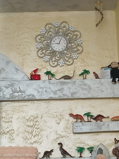 The image depicts a shelf display featuring various decorative items and figurines. At the top, a decorative wall clock with an ornate silver frame and crystal embellishments is visible. To its right, a small, hanging chandelier-like ornament with clear beads and dark gray accents is suspended from the ceiling.

On the first shelf from the top, a small Santa Claus figurine in red attire with a white beard is placed next to a red bag. To its right are two miniature palm trees. Further to the right, a plastic dinosaur figurine in a reddish-brown color stands next to a white box. A stuffed monkey with a brown body and lighter face is positioned on the far right of this shelf, partially obscuring the box. A long-necked dinosaur figurine is on the left side of the shelf.

The second shelf from the top displays several plastic dinosaur figurines. On the left, a red Tyrannosaurus Rex is positioned next to two green palm tree figurines. To the right of the palm trees is a brown Stegosaurus figurine, followed by a light brown quadrupedal dinosaur.

The third shelf from the top shows a dark gray dinosaur figurine on the left, standing next to two green palm tree figurines. In the center of this shelf, a white oscillating fan with a mesh cover is visible. Part of a stuffed animal with a leopard-like pattern is visible on the far right.

The background wall has a textured, off-white or cream color with darker vertical streaks. The shelves themselves appear to be made of wood or a similar material, painted in a textured white and gray finish. The image appears to be taken indoors. The lower left corner of the image shows a watermark indicating "nubia Focus 2 Ultra 5G".

There is no information available in the image to confirm its location in Samuilovo, Bulgaria.