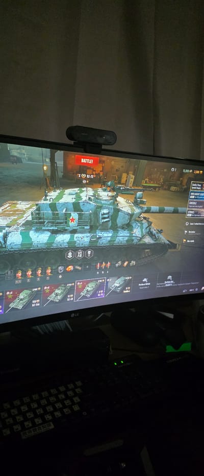 The image shows a computer monitor displaying the game "World of Tanks." The game features a garage view with a tank, identified as the BZ-75, prominently displayed. Various game interface elements are visible, including battle status, vehicle statistics, and in-game currency. The monitor is positioned on a desk, with a keyboard and a computer mouse visible in the foreground. A webcam is mounted on top of the monitor. The environment appears to be a dimly lit room, possibly a home office or gaming setup. The image was captured in Gimhae-si, South Korea.