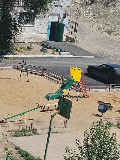 The image depicts a children's playground located in Jezqazğan, Kazakhstan, in front of a drab, multi-story apartment building. The playground area is covered with sand and features several pieces of equipment. In the foreground, a green seesaw with yellow supports is visible. To the right of the seesaw, a blue and yellow spring rider shaped like a helicopter is situated. Behind these, a green and yellow slide with red ladder rungs stands prominently. A metal swing set with a dark frame is positioned further back. The apartment building in the background is weathered, with cracked concrete walls and a green entranceway. Tires are stacked and used as planters near the building's entrance. A dark-colored sedan is parked on the asphalt area adjacent to the playground. The overall environment is dusty and sparsely vegetated, typical of an urban setting in a dry climate.