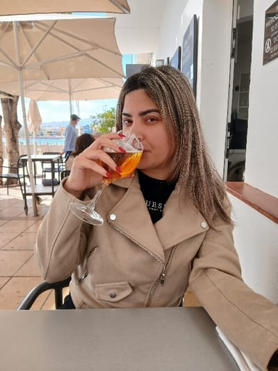 The image features a young woman seated outdoors at a cafe or restaurant in Agüimes, Spain, during the daytime. She has long, straight hair with light brown and blonde highlights and is looking directly at the camera with a calm expression. She is wearing a light beige, suede-like biker jacket with silver zippers and stud details, over a dark top that visibly displays the "GUESS" logo. Her fingernails are painted red.

She is holding a stemmed glass to her lips, in the act of drinking an orange-colored beverage, likely a cocktail such as an Aperol Spritz, with visible ice cubes. The setting appears to be an open-air terrace or patio. A light-toned table is in front of her. In the background, multiple large, light-beige patio umbrellas provide shade, indicating bright, sunny weather. Beyond these umbrellas, there is a picturesque view of a body of water—likely the sea or a bay—with distant buildings or a town visible along the shoreline, consistent with a coastal location. Another individual is partially visible in the mid-ground, walking away. Dark signs with white text are mounted on a wall in the far background, but the text is illegible. The overall scene suggests a relaxed atmosphere in a pleasant outdoor environment.
