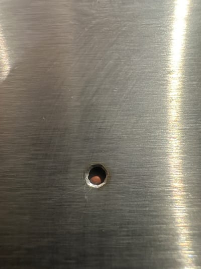 This image is a close-up, eye-level shot of a brushed metal surface, likely stainless steel. The surface has a matte finish with visible linear brush strokes that run vertically from top to bottom. A bright light source is reflected off the upper portion of the surface, creating a strong, blown-out highlight with subtle rainbow-like iridescence, indicating the metallic nature and reflective quality of the material.

The central subject of the image is a small, circular hole in the metal. The edges of the hole appear slightly rough and irregular, suggesting it may have been drilled or punched. Through the hole, a glimpse of a coppery or reddish-brown material can be seen, possibly the material behind the metal sheet or a fastener embedded within it.

The overall scene is minimalist and industrial, focusing solely on the texture and details of the metal and the hole. There are no people, animals, or explicit environmental cues to suggest a specific time of day or weather conditions. The lighting is artificial, given the harsh reflection. The location context of Letterkenny, Ireland, does not provide any direct visual clues within the image itself, as the metal surface is generic. No text is visible in the image. The focus is on the tactile and visual qualities of the damaged metal surface.