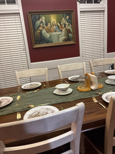 A dining room features a long, wooden table meticulously set with ornate plates, gold cutlery, and a green runner. The table is flanked by white chairs. On the wall, a framed painting depicts a religious scene, possibly the Last Supper. Blinds cover the windows above and below the painting, creating a contrast with the dark red wall.