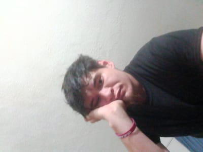 A man is lying down with his head resting on his left hand, which has a pink bracelet. He is wearing a black shirt. The background is a light-colored wall. Location information is not discernible from the image.