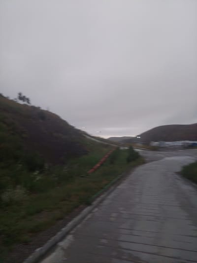 The image depicts a gray, overcast sky above a landscape that includes a steep, grassy hill on the left and a paved road curving towards the right. Sparse vegetation grows on the hillside, and what appear to be distant lights are visible on the horizon. Structures, possibly industrial buildings, are partially visible in the middle ground. The road appears wet, suggesting recent rain. The scene has a muted, somber atmosphere. The location is Bordunskiy, Kyrgyzstan.
