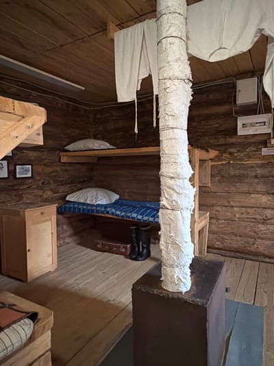 The image depicts the interior of a rustic wooden room, likely a cabin or dwelling in Ust'-Pyzas, Russia. The room is sparsely furnished, suggesting a functional living space rather than a display of luxury. The walls and ceiling are constructed from rough-hewn logs, giving the space a natural and aged appearance.
Dominating the scene are bunk beds, indicating shared living quarters. The lower bunk is made up with a blue and white checkered mattress and a white pillow. On the floor beneath the lower bunk, a brown suitcase and a pair of black boots are placed, suggesting recent occupancy or preparation for travel. The upper bunk, also with a white pillow, is simpler in its arrangement. White fabric, possibly clothing or linens, is hung from the ceiling to the right of a central, vertically oriented pipe. This pipe is wrapped in what appears to be insulating material, secured by thin wires.
To the left of the bunks, a small, light-colored wooden cabinet stands with a single drawer and a door, implying storage space. Above the cabinet, two framed pictures are hung on the log wall, adding a personal touch to the otherwise spartan environment.
The floor is made of wooden planks. In the foreground, partially visible, is a low wooden structure with a patterned cushion, possibly another seating or sleeping surface. A dark metal object, potentially a stove or furnace, is positioned in the right foreground, with the insulated pipe rising from it. Electrical outlets and a distribution box are visible on the log wall in the background, indicating some degree of modern utility within the rustic setting. The overall impression is one of a simple, practical living space, possibly in a remote or historical context.