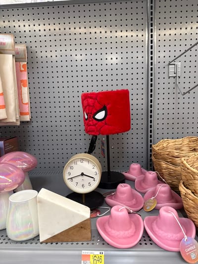 The image captures a retail store display, rotated 90 degrees clockwise, showcasing various home decor and gift items against a grey pegboard wall. In the center, a red, fuzzy cube-shaped item adorned with a Spider-Man mask design is mounted on a black stand. To its right, an analog clock with a white face, black numbers, and hands, showing approximately 2:40, is also displayed on a black stand.

Above the clock and Spider-Man item, several pink, cowboy hat-shaped candles are hanging from display hooks. These candles have white tags that read "CANDLE" and "HAT CANDLE." A yellow price tag nearby indicates "$646," likely representing $6.46, and also reads "HAT CANDLE." Towards the top of the frame, a textured, light brown woven basket or decorative item is visible.

Below the clock, a white, triangular object, possibly a decorative piece made of stone or ceramic, rests next to a wooden block. On a lower shelf, iridescent pink egg-shaped objects and clear or white iridescent cylindrical vases or cups are displayed. Further down, stacks of folded fabric or blankets with pink and white patterns are arranged on a shelf. The pegboard wall features numerous small holes, and several metal hooks are visible, some empty and some holding items. No people are present in the image. Partial text like "AW" and "UDAH" is also visible in the OCR.