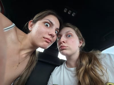 Two young women are inside a car. Both are making exaggerated facial expressions with wide eyes and pursed lips. The woman on the left has dark hair, a gold necklace, and is wearing a white top with a white strap visible. The woman on the right has light brown hair and is wearing a white t-shirt. The car's interior includes black seats and overhead lights. The location is unidentifiable as Chokoba, Bulgaria from the given image.
