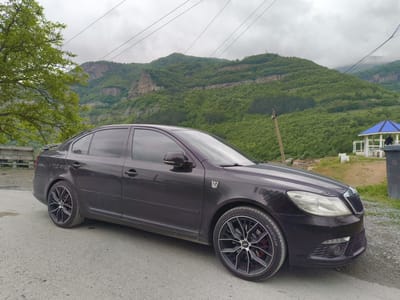 A dark-colored Skoda Octavia sedan is parked on a narrow asphalt road. The car features aftermarket black wheels with a spoke design and red brake calipers. A Transformers Autobot emblem is affixed to the front fender. The backdrop consists of a verdant mountain landscape with layered rock formations under an overcast sky. Utility poles and wires are visible overhead. To the right of the car, a white gazebo with a blue roof stands on a grassy area. A waste bin marked with the number 68 is nearby. The setting appears to be in the vicinity of Bov, Bulgaria, based on the terrain and architectural elements.