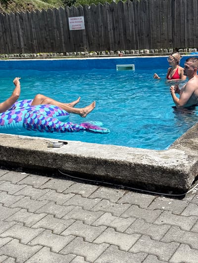 In Liava Reka, Bulgaria, a group of four individuals are in an outdoor swimming pool on a sunny day. The pool has clear blue water and is surrounded by a concrete edge and a grey paver pathway. A wooden fence runs along the back of the pool area, with a sign in Bulgarian attached to it. One person is partially submerged in the water, floating on a colorful inflatable shaped like a fish tail. Another person, wearing a red bikini, is also in the water, interacting with a male figure who is mostly out of frame. A pack of cigarettes and a lighter are placed on the concrete edge of the pool. The atmosphere appears relaxed, with people enjoying a leisurely day by the water.